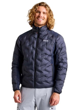 Un hombre sonriente de pelo largo y rizado luce una chaqueta acolchada azul marino con cremallera y unos pantalones gris claro sobre un fondo blanco.