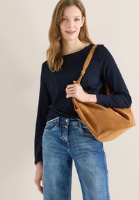 Femme portant un haut noir à manches longues et un jean bleu, tenant un grand sac à bandoulière en daim marron sur un fond beige.