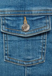 Detail kapsy z džínoviny s světle modrou tkaninou, oranžovým švem a stříbrným zapínáním s embosovaným textem, který ukazuje texturovaný povrch.