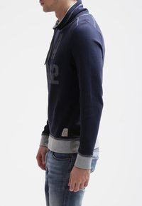 Felpa con cappuccio blu navy e dettagli grigi, realizzata in tessuto morbido. Presenta una stampa frontale e un cappuccio con coulisse. Polsini e orlo a costine.