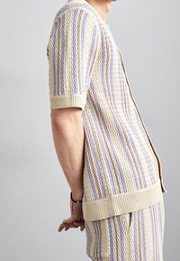 Gebreid vest in crème, met paarse en lavendelkleurige strepen. Korte mouwen, geribbelde manchetten en zoom, gestructureerd patroon en open voorkant.