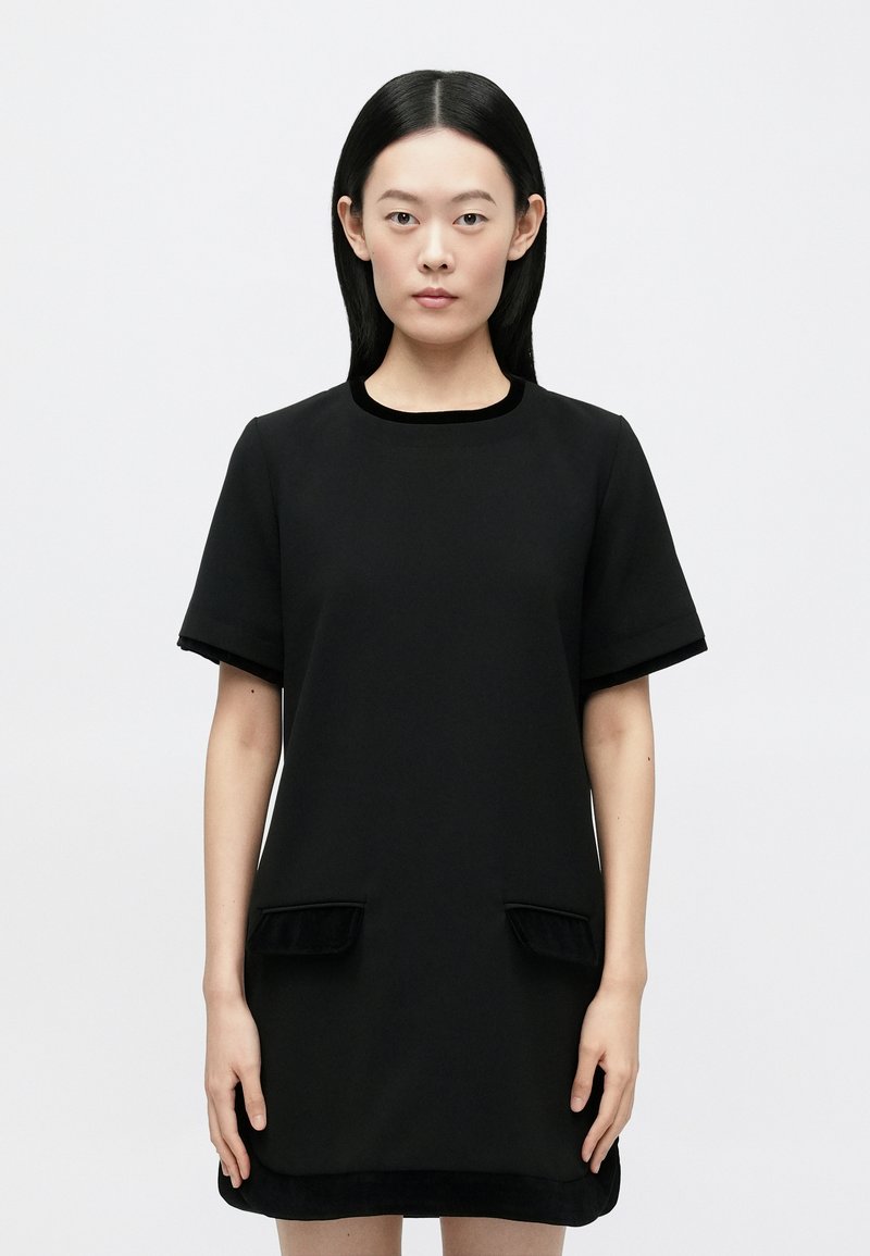 Mujer con cabello liso y negro, vestida con un vestido negro de manga corta con dos bolsillos delanteros, de pie contra un fondo claro y liso.