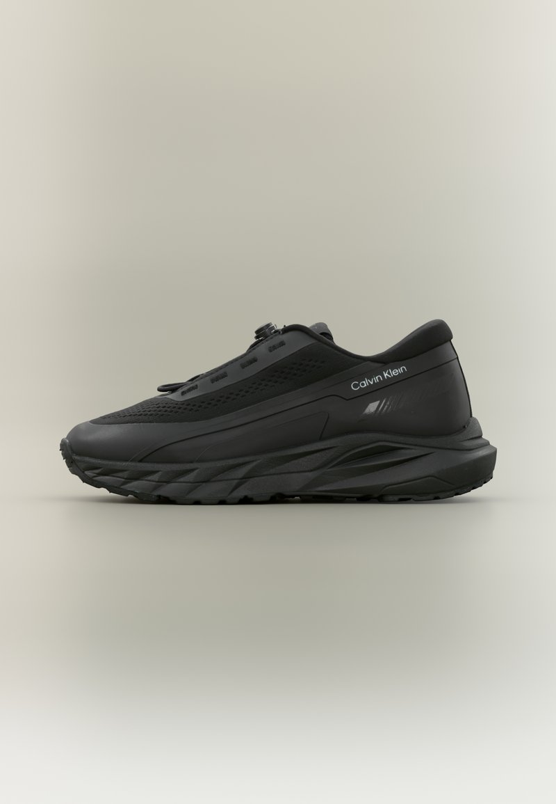Μαύρο αθλητικό παπούτσι Calvin Klein με παχιά σόλα, ανώτερη λεπτομέρεια από δίχτυ, ρυθμιζόμενο κορδόνι με διακόπτη και λογότυπο στην πλευρά, εμφανίζεται σε αριστερή όψη.