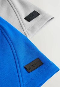 Due maniche di tessuto, una grigia e una blu. Entrambe presentano un'etichetta nera testurizzata con un logo, mostrando una superficie liscia e lavorata a maglia.