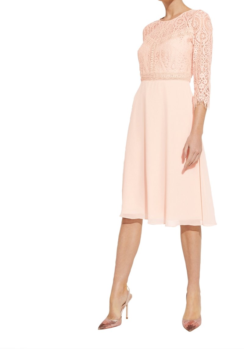 Femme portant une robe rose clair longueur genou avec des manches trois-quarts en dentelle et des chaussures à talons hauts à bout pointu.