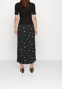 Jupe maxi noire à motifs floraux jaunes et turquoises, associée à un haut noir ajusté, portée avec des sandales à talons blocs.