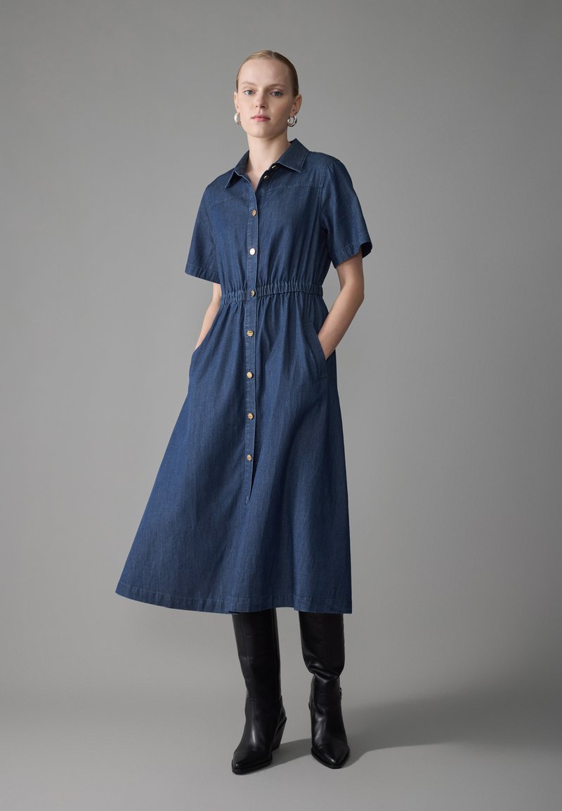Denim knappkjole med korte ermer, innsnørt midje og lommer. Har en full skjørt, naturlig fibert tekstur, og gullknapper.