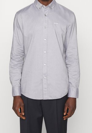 Mężczyzna w jasnoszarej koszuli zapinanej na guziki z długim rękawem i logo "BOSS" na lewej piersi, w połączeniu z ciemnymi spodniami, stoi na jednolitym tle.