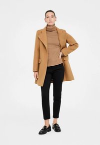 Zandkleurige jas van een wolmix met een reverskraag, gecombineerd met een bruine geribbelde coltrui, zwarte nauwsluitende broek en zwarte loafers. Minimalistisch ontwerp.