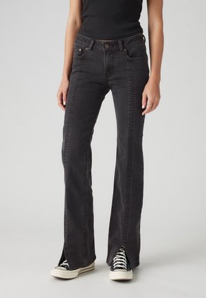 Personne portant un jean flare noir avec des détails de couture à l'avant et des fentes aux chevilles à l'avant, associé à des baskets noires et blanches.