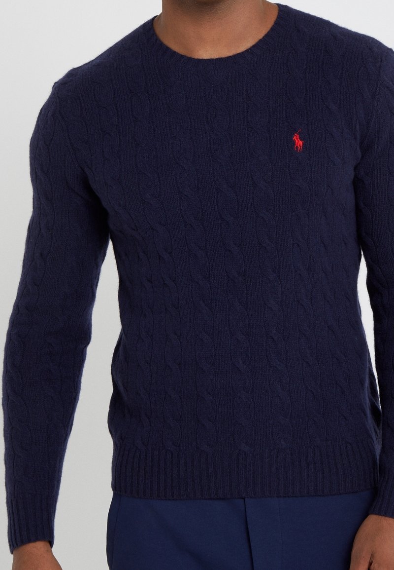 Pull en maille torsadée bleu marine avec logo brodé d'un joueur de polo rouge sur la poitrine gauche, porté par une personne à la peau foncée.