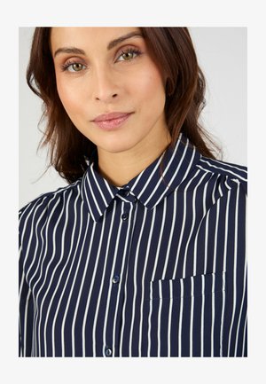 Junge Frau mit braunen Haaren, die ein dunkelblaues Hemd mit weißen vertikalen Streifen und einer Fronttasche trägt, leicht lächelnd vor einem schlichten Hintergrund.