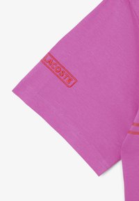 Manica di t-shirt in cotone rosa con etichetta ricamata "LACOSTE" arancione, caratterizzata da sottili righe orizzontali di un colore coordinato.
