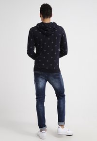 Černá mikina s hvězdným vzorem, měkká tkanina, kapuce na šňůrku, kombinovaná s modrými džíny a bílými teniskami, pohled zezadu.