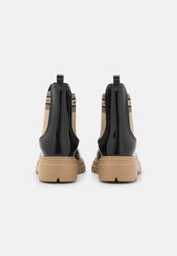 Botines negros brillantes con paneles de lona beige, con una suela gruesa y texturizada en tono marrón claro y lengüetas traseras para facilitar su puesta.