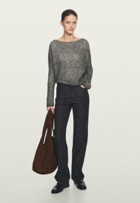 Donna che indossa un maglione grigio a maglia, jeans neri e scarpe nere, mentre tiene una grande borsa a tracolla marrone con una mano in tasca.