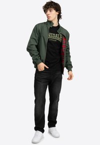Chaqueta bomber verde con forro de tartán, camiseta negra con letras blancas, jeans negros y zapatillas blancas. Ajuste informal, tejido suave.