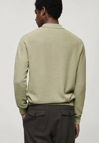Long-sleeve polo shirt in light green textured knit, featuring a folded collar and a relaxed fit. Paired with dark brown trousers.
