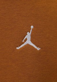 Tessuto marrone con un logo ricamato bianco raffigurante una figura stilizzata che salta per tirare a canestro, su uno sfondo a maglia strutturata.