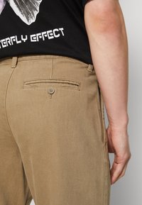 Chinos beige con una fabbrica texturizzata, design aderente e una tasca posteriore con chiusura a bottone, con una mano parzialmente visibile appoggiata sui fianchi.