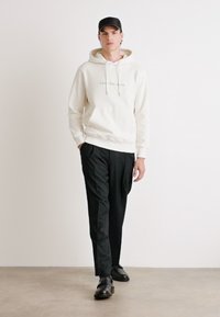 Sudadera crema clara con cordón, que presenta texto, combinada con pantalones negro ajustados y zapatos negros brillantes. El modelo se encuentra sobre un fondo neutro.