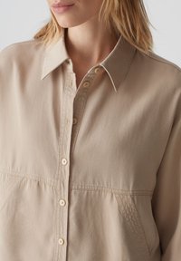Beige button-up overhemd met een kraag, voorzien van een gestructureerde stof, verticale naden en lichtbruine knopen langs de voorflap.