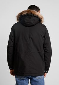Parka negra con capucha forrada de piel, que presenta una textura suave y un diseño recto. Incluye costuras visibles y botones a presión en el dobladillo.