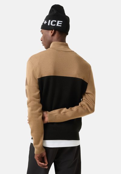 Strikket pullover i tan og sort med høj krave; har struktureret stof, lange ærmer og et geometrisk design på overarmen.