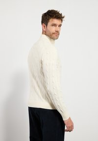 Cremfarbener Kabelstrickpullover mit hohem Kragen, gerippten Bündchen und Saum. Getragen mit dunklen Cordhosen. Klare Textur- und Musterdetails sichtbar.