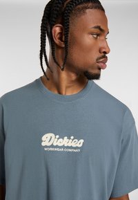 Puuvillainen t-paita liilassa sinisessä, jossa on teksturoitu kerma logo, jossa on brodeerattu "Dickies Workwear Company" edessä. Pyöreä kaula-aukko.
