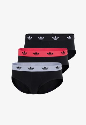 Jonge man met sproeten en dreadlocks, staand op blote voeten, dragend een zwarte slip met een witte tailleband met een zwart trefoil-logo.