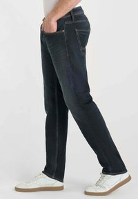 Jean en denim foncé avec une coupe droite, dotés de coutures orange, de poches latérales et d'un léger brillant. Portés avec des baskets blanches.