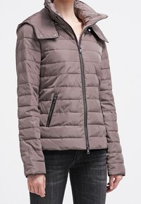 Padded jacket w kolorze jasnobrązowym z wysokim kołnierzem i klapami na ramionach zapinanymi na zatrzaski. Posiada zamek błyskawiczny w przednich kieszeniach i zamek błyskawiczny na środku.