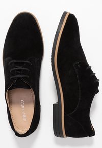 Chaussures oxford en suède noir avec une semelle plate, un laçage à l'avant et une accent en tan autour de la semelle. La doublure intérieure est beige clair.