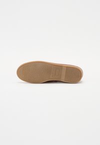 Zapatilla marrón con parte superior suave, suela de goma plana, fondo texturizado para agarre y diseño minimalista. Sin logotipos ni acentos visibles.