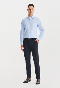 Mężczyzna w jasnoniebieskiej koszuli z guzikami, ciemnych dopasowanych spodniach oraz brązowych eleganckich butach, stoi z jedną ręką w kieszeni.