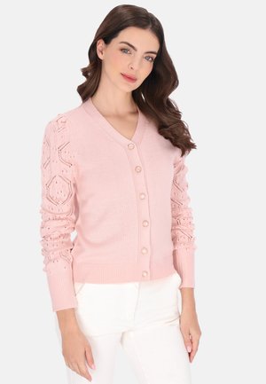 Světle růžový pletený kardigan s výstřihem do V, zapínáním na knoflíky a vzorovanými rukávy. Má žebrované manžety a lem. Výrazný krajkový detail na rukávech.
