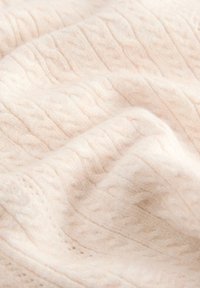 Tessuto beige chiaro con una texture morbida che presenta un motivo a treccia lavorata a maglia in rilievo. La superficie appare soffice e leggermente ondulata.