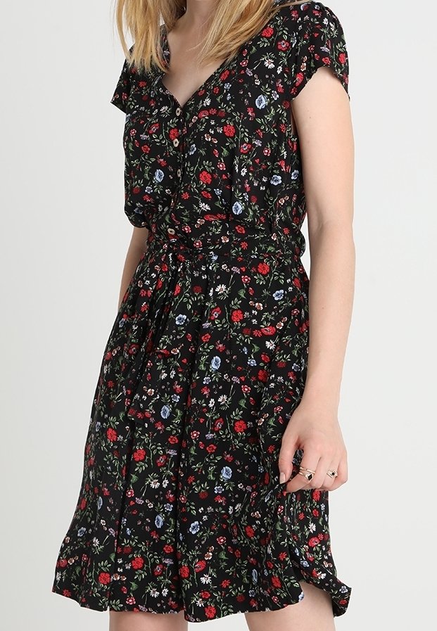 Femme portant une robe noire à motifs floraux rouges, blancs et bleus, manches courtes, boutons sur le devant et une ceinture à la taille.