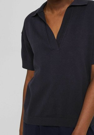 Osoba ubrana w ciemnogranatową, krótkorękawową, dzianinową koszulkę polo z rozpiętym kołnierzykiem typu V, na jednolitym tle.