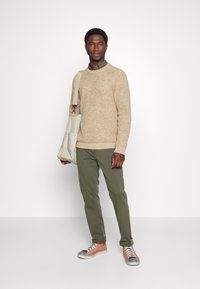Maglione beige con texture, camicia a quadri, pantaloni verde oliva e sneakers arancioni con accenti grigi. Porta una borsa a tracolla beige.