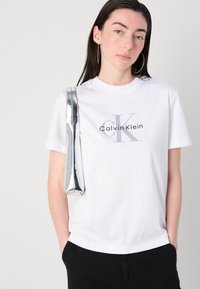 Camiseta blanca de algodón con un logo gris de "Calvin Klein", combinada con un bolso metálico plateado. El modelo tiene el cabello largo y lleva pendientes de aro.