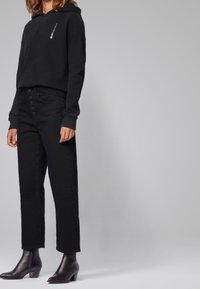 Svart huvtröja med vitt logotyp, kombinerad med högmidjade svarta jeans med knappspärr och svarta klackade ankelskår.