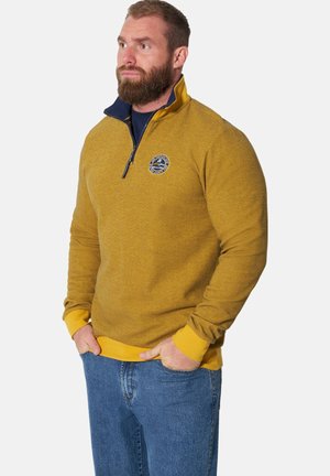 Man with beard wearing mustard yellow quarter-zip sweater with logo and blue jeans, standing with hands in pockets, looking left.