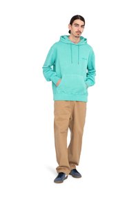 Sudadera turquesa con bolsillo frontal y cordones, combinada con pantalones beige y zapatillas marinas. Textura suave y corte relajado.