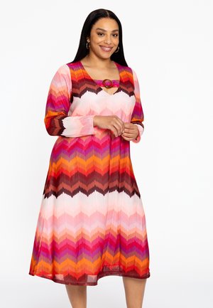 Robe à motif chevron coloré en rose, orange et marron, avec un décolleté en V et un accent décoratif rond au niveau de la poitrine. Manches longues.