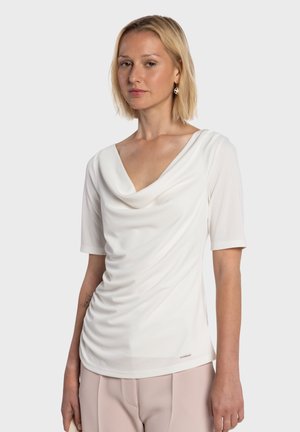 Haut blanc à col cheminée avec manches courtes, texture douce et détails froncés sur le devant, associé à un pantalon de couleur claire.