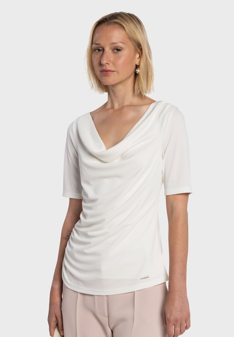 Top bianco con collo a fodero, maniche corte, tessuto morbido e dettagli arricciati sul davanti, abbinato a pantaloni chiari.