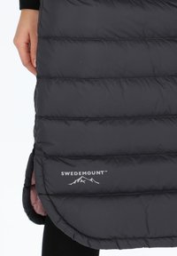 Musta untuvapuku, jossa on ommellut kammiot ja korkean matalan helma. Vahvistettuna hopeavärisellä "SWEDEMOUNT" -logolla, jossa on vuoristokuvasto. Vuorattu kontrasti-pinkillä.