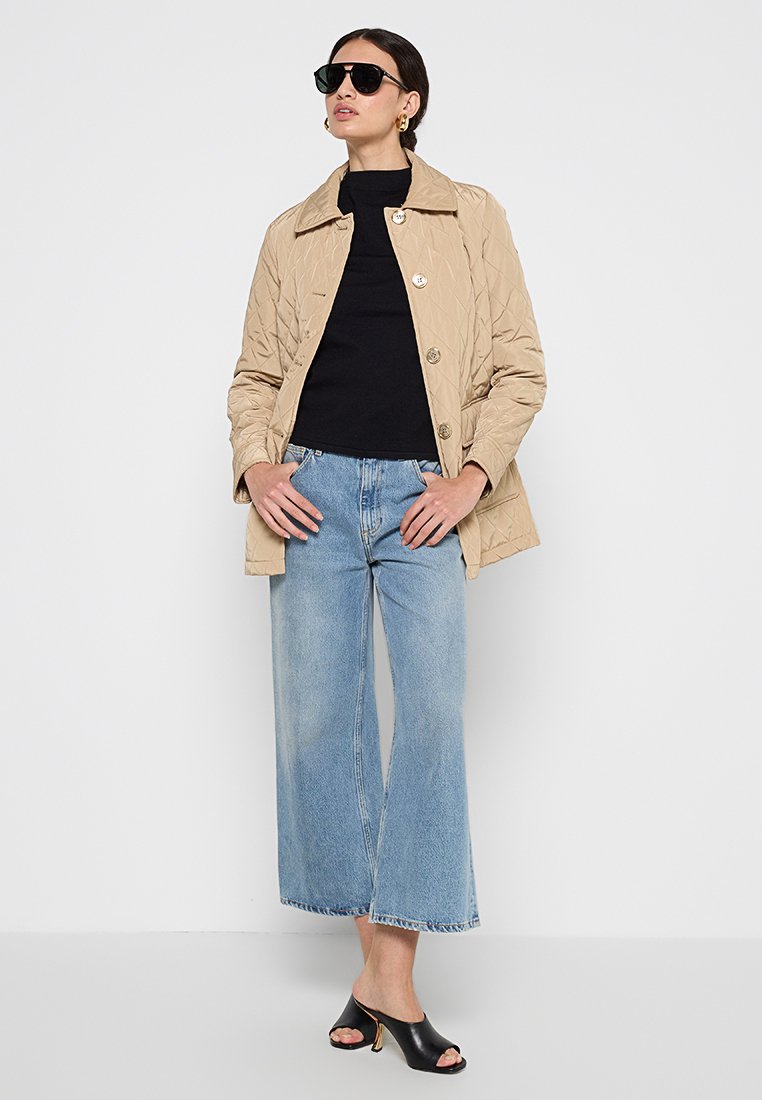 Giacca trapuntata beige con bottoni grandi, indossata sopra una maglietta nera, abbinata a jeans a gamba larga azzurri e mule neri con punta aperta.